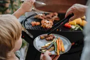 Grå tallerken med grillede grøntsager og kød, herunder gulerødder, squash og pølser, serveres fra udendørs grill. Barn holder tallerken, voksen bruger grilltang.