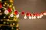 Lyskæde med runde hvide pærer og røde pomponer, festlig dekoration, sløret juletræ med gule lys i baggrunden, velegnet til jul eller fest.