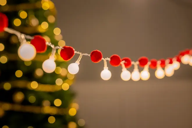 Lyskæde med runde hvide pærer og røde pomponer, festlig dekoration, sløret juletræ med gule lys i baggrunden, velegnet til jul eller fest.
