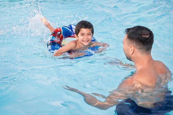 Barn simmar i pool med blå badring med Spiderman-motiv, övervakad av vuxen man i vattnet. Badringen ger flythjälp och stöd vid simning.
