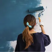 Kvinde maler væg med blå maling og rulle. Hun har mørk skjorte, hvid handske og blå sløjfe i håret. Produkt: blå vægmaling til indendørs brug.