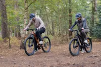 Två vuxna cyklar på mountainbikes med tjocka däck, framdämpning och skivbromsar. Båda cyklar har raka styren och passar för terräng. Hjälmar och fritidskläder används.
