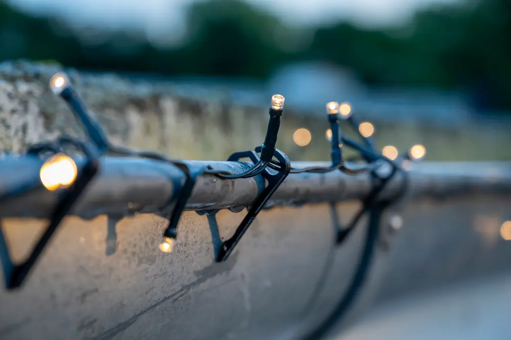Udendørs lyskæde med små, varm-hvide LED-pærer på sort ledning, fastgjort til en betonoverflade. Perfekt til dekorativ belysning på terrasser, altaner eller i haven.