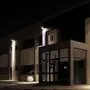 Moderne bygningsindgang med store glasdøre og vinduer, oplyst af udendørs væglamper. Minimalistisk design med hvid plantekrukke og plante ved indgangen. Nattesetting.