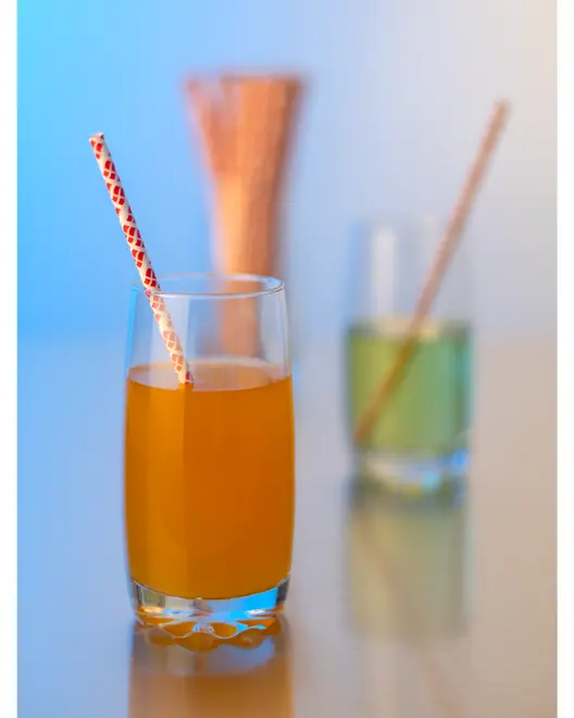 Høj klar glas med appelsinjuice og dekorativt papirsugerør. Moderne, minimalistisk design til kolde drikke. Baggrunden viser et andet glas med grøn væske og sugerør.