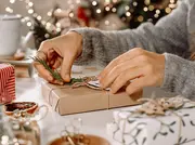 Händer slår in en present i brunt papper, knyter med snöre och dekorerar med en grön kvist för en rustik, miljövänlig julklapp.