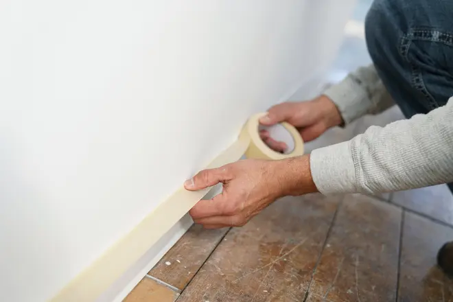 Person påfører malertape langs bunden af en hvid væg for at beskytte gulvet før maling. Tapen er beige, gulvet er brune fliser, personen har grå trøje på.