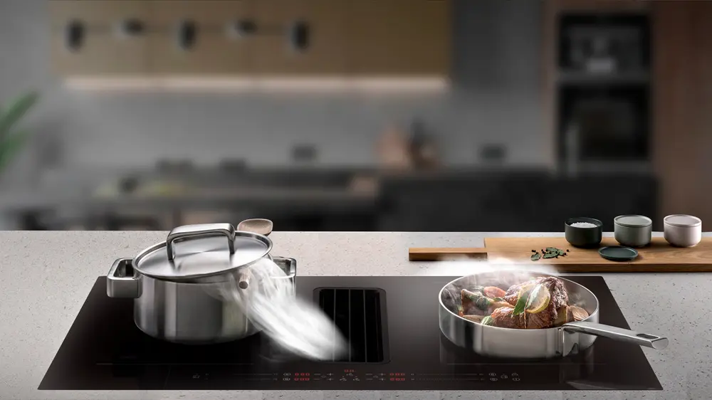Modern induktionshäll med integrerad fläkt, rostfri gryta och stekpanna. Grytan ångar, pannan innehåller kött och grönsaker. Snygg svart glasyta.