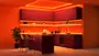 Moderne køkken med bordeaux skabe, orange LED-belysning, køkkenø med to barstole, indbyggede apparater, åbne hylder og en potteplante. Elegant, farverig indretning.