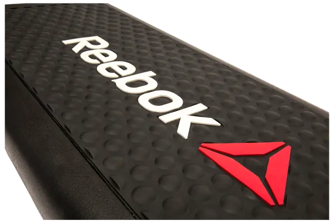 Sort Reebok stepbænk med skridsikker, nopret overflade, hvidt Reebok-logo og rød trekantet emblem. Robust plast, ideel til aerobic og steptræning.