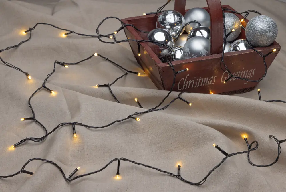 A string of warm white LED fairy lights with a black wire is spread out on a beige fabric surface. The lights are evenly spaced and emit a soft glow. In the background, there is a rustic wooden basket with the words 'Christmas Greetings' engraved on the side, filled with shiny silver and metallic Christmas baubles in various finishes, including matte, glossy, and glittery.