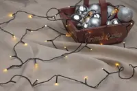 A string of warm white LED fairy lights with a black wire is spread out on a beige fabric surface. The lights are evenly spaced and emit a soft glow. In the background, there is a rustic wooden basket with 'Christmas Greetings' written on the side, filled with shiny silver and metallic Christmas ornaments in various finishes, including matte, glossy, and glittery textures.