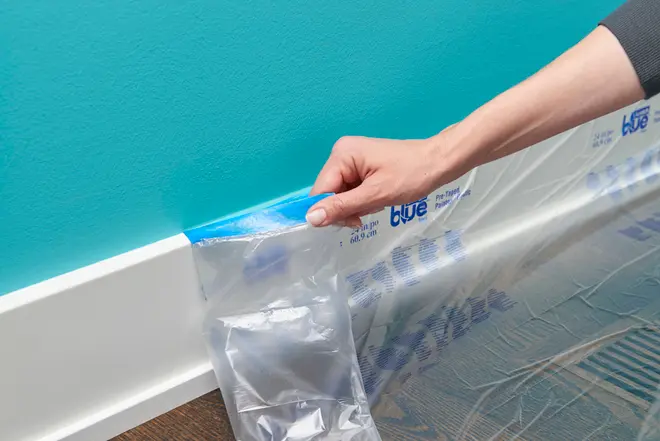 Beskyttende plastfolie med blå malertape fastgjort, påført langs en hvid fodliste for at beskytte gulv og væg under maling. Perfekt til præcise maleropgaver og nem fjernelse.