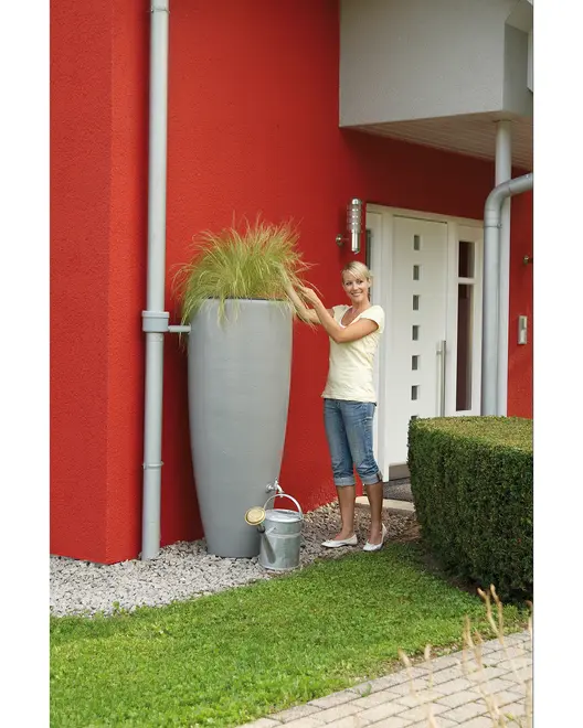 Høj, moderne grå regnvandstønde med integreret plantekasse øverst, placeret op ad husmur, har vandhane for nem adgang og vandkande under.