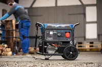 Bærbar generator med hjul og håndtag, flere strømudtag, kontrolpanel og robust ramme. Velegnet til udendørs eller nødbrug, vist i et værkstedsmiljø.