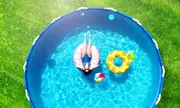 Rund ovanmarkspool med blå liner, inkluderar uppblåsbar donut och ananasbadring samt en färgglad strandboll. Poolen står på gräs, perfekt för sommaren.