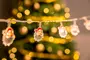 Lyskæde med små julemandsansigter med røde hatte, ideel til juledekoration. Baggrunden viser varme gule bokeh-lys og et sløret juletræ.