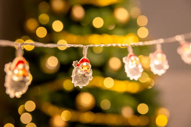 Lyskæde med små julemandsansigter med røde hatte, ideel til juledekoration. Baggrunden viser varme gule bokeh-lys og et sløret juletræ.