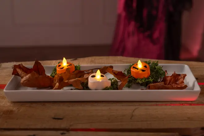 Rektangulär vit bricka med tre LED-värmeljus formade som pumpor och en dödskalle, omgivna av torkade löv och grön mossa, perfekt för Halloween-dekoration.