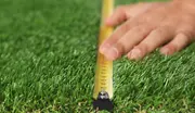Nærbillede af en hånd, der måler kunstgræs med et gult målebånd, hvilket viser det realistiske grønne udseende og tætheden af det syntetiske græs.