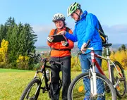 To personer med hjelme og udendørsjakker står med mountainbikes og bruger en tablet. Cyklerne har affjedring foran, grove dæk og drikkedunke, velegnet til terrænkørsel.