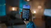 En hånd holder en smartphone med en smart home-app, der viser styring af stue, køkken, spisestue og soveværelse. Sløret baggrund af et hjem.
