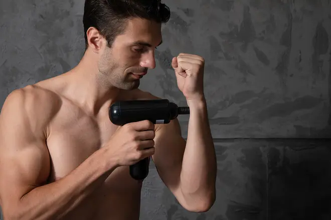 Håndholdt sort massagepistol med ergonomisk greb, bruges på underarmsmuskler. Har cylindrisk krop, afrundet massagehoved og er designet til dybdegående muskelafslapning og restitution.