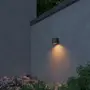 Moderne udendørs væglampe med cylindrisk sort armatur, monteret på grå væg, udsender varmt lys nedad. Perfekt til belysning af have eller terrasse.