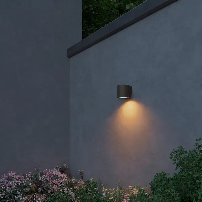 Moderne udendørs væglampe med cylindrisk sort armatur, monteret på grå væg, udsender varmt lys nedad. Perfekt til belysning af have eller terrasse.