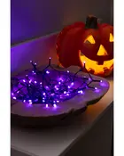 Lila LED-ljusslinga i en träskål, lyser starkt. I bakgrunden syns en upplyst orange pumpalykta för Halloween.
