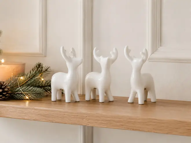 Tre blanke, hvide keramiske rensdyrfigurer står på en træhylde, stylet med varm julegrønt og lys. - Billedet er skabt med AI