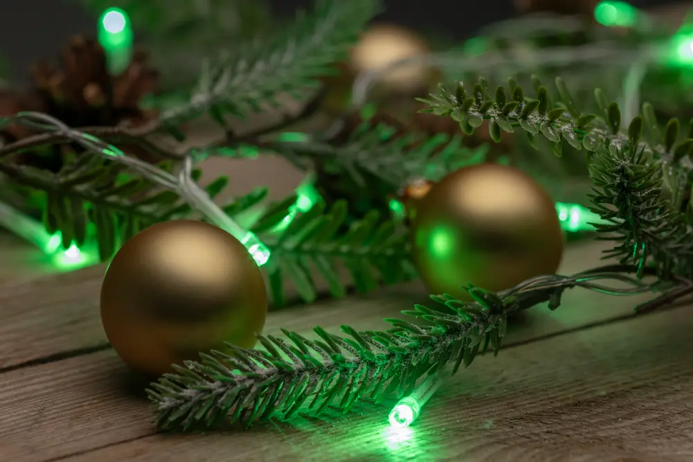 Konstgjorda grankvistar med gröna LED-ljusslingor och matta guldfärgade julkulor, arrangerade på en träyta för festlig dekoration. Kottar syns i bakgrunden.