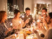 Festligt dukat middagsbord med olika maträtter, levande ljus, vinglas och personer som håller tomtebloss. Mysig inomhusmiljö med varm belysning och ljusslingor i bakgrunden.