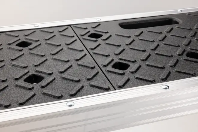 Närbild av svart, halkfri plattform med diamantmönster, fyrkantiga utskärningar och handtag, inramad av silverfärgad metall med synliga skruvar.