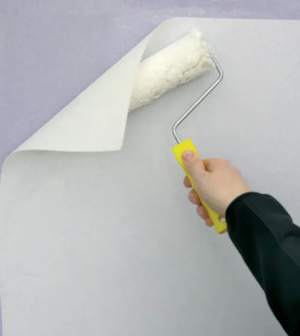 En person bruger en malerrulle med gult håndtag til at påføre lim eller maling på en væg, klar til at opsætte eller udjævne hvidt tapet.