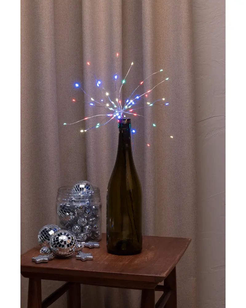 Vinflaska med flerfärgade LED-ljusslingor arrangerade som fyrverkerier, används som dekorativ mittpunkt. Glasburk med små discobollar och stjärnformade dekorationer intill.