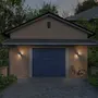 Moderne garage med blå sektionalport, udendørs væglamper, beige vægge, tegltag og cykel parkeret til højre på indkørslen. Omgivet af grønne planter.