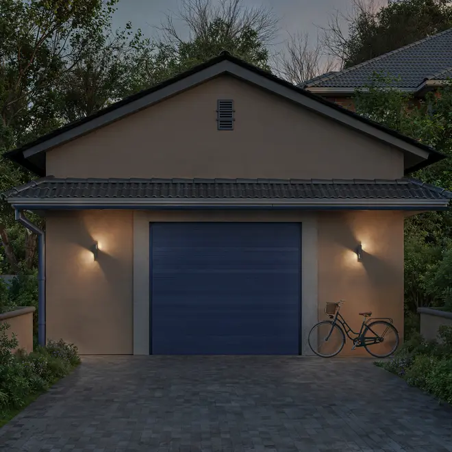 Moderne garage med blå sektionalport, udendørs væglamper, beige vægge, tegltag og cykel parkeret til højre på indkørslen. Omgivet af grønne planter.