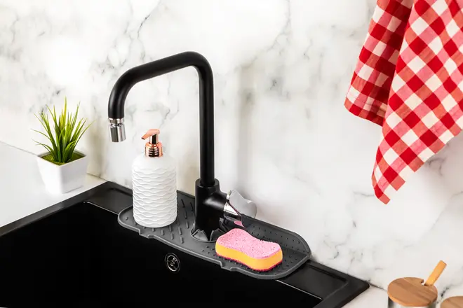 Sort silikone måtte til vandhane, passer omkring vaskens armatur. Holder sæbedispenser og pink svamp. Beskytter bordpladen mod vand og organiserer rengøringsartikler.