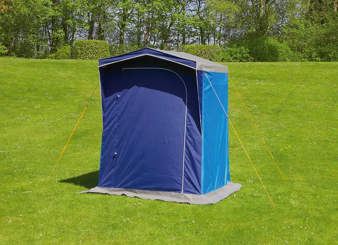 Bærbart blåt privattelt med spidst tag, lynlåsdør foran og barduner for stabilitet. Velegnet til campingbad eller omklædning. Står på græsplæne udendørs.