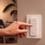 En hånd trykker på en hvid Philips Hue smart-lyskontakt med fire knapper til strøm, lysstyrkejustering og hue-kontrol, monteret på en væg.
