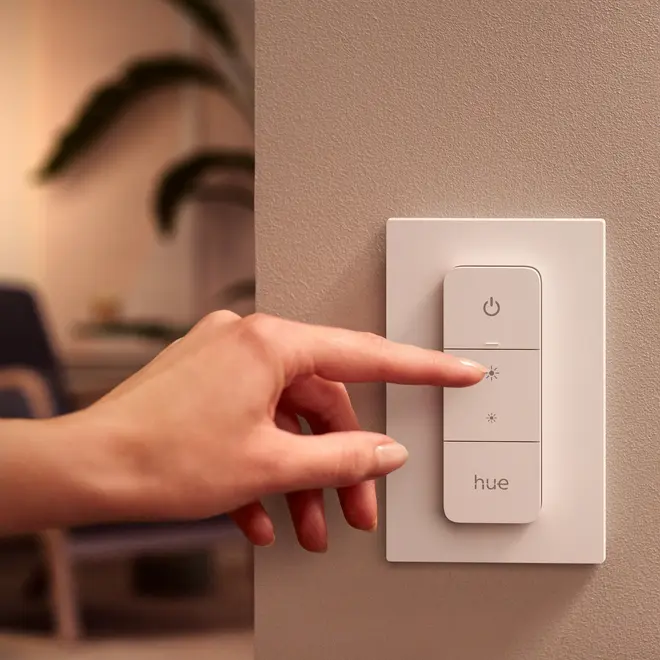 En hånd trykker på en hvid Philips Hue smart-lyskontakt med fire knapper til strøm, lysstyrkejustering og hue-kontrol, monteret på en væg.