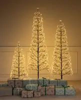 Three minimalist artificial trees of varying heights, each with bare branches adorned with numerous warm white LED lights, creating a glowing effect. The trees have a slender, modern design and are displayed indoors against a plain wall. At the base, there are several wrapped gifts in pastel and neutral tones, featuring various patterns and tied with green or white ribbons.