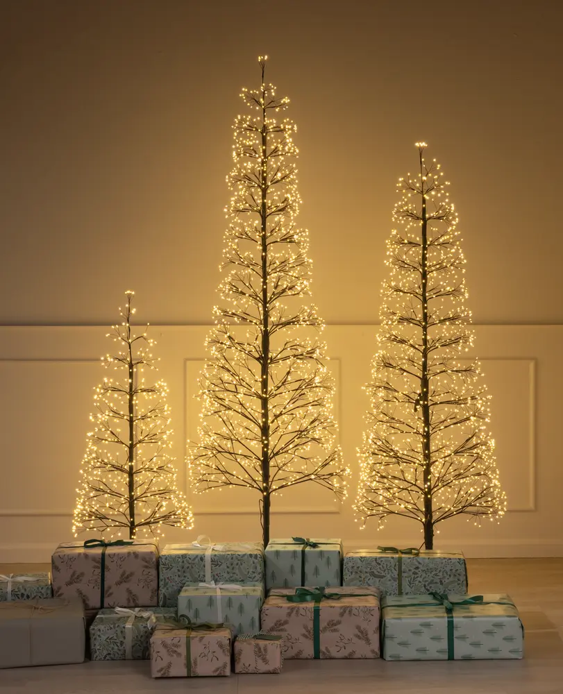 Three minimalist artificial trees of varying heights, each with bare branches adorned with numerous warm white LED lights, creating a glowing effect. The trees have a slender, modern design and are displayed indoors against a plain wall. At the base, there are several wrapped gifts in pastel and neutral tones, featuring various patterns and tied with green or white ribbons.