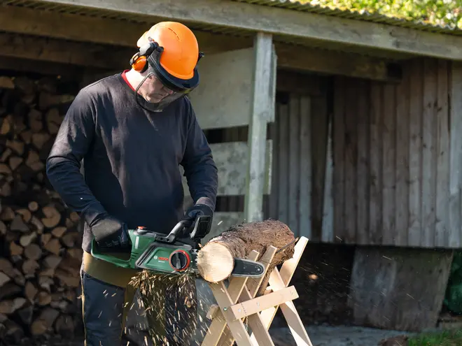 Elektrisk kædesav med grønt hus, sort klinge og sikkerhedsfunktioner. Bruges til at save brænde, vist med beskyttelseshandsker og hjelm. Velegnet til havearbejde og træskæring.