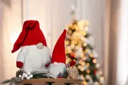 Två mjuka juldekorationstomtar med röda mössor, vita skägg och runda näsor, dekorerade med kottar och grankvistar, perfekta för julpynt i hemmet.
