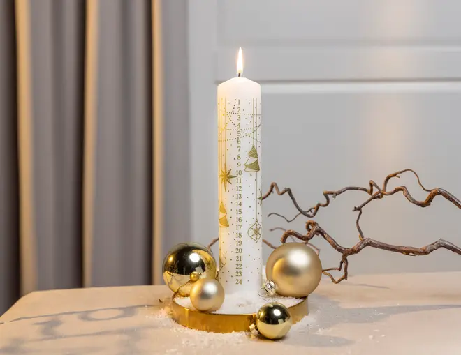 Vit kalenderljus med guldfärgade julmotiv och siffror, omgiven av guldkulor på träbricka. Dekorativ gren och snölika detaljer ger en festlig känsla.