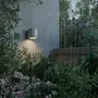 Moderne cylindrisk udendørs væglampe i mat grå, monteret på væg, lyser haveområde op. Vejrbestandigt design, vist i regn, egnet til udendørs brug.