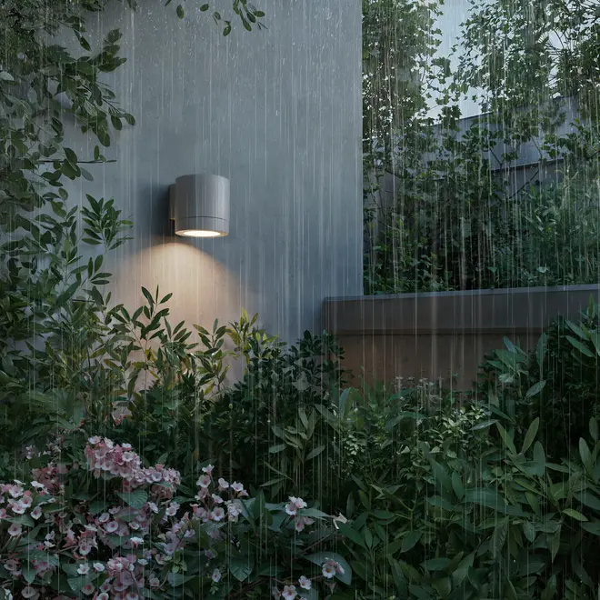 Moderne cylindrisk udendørs væglampe i mat grå, monteret på væg, lyser haveområde op. Vejrbestandigt design, vist i regn, egnet til udendørs brug.