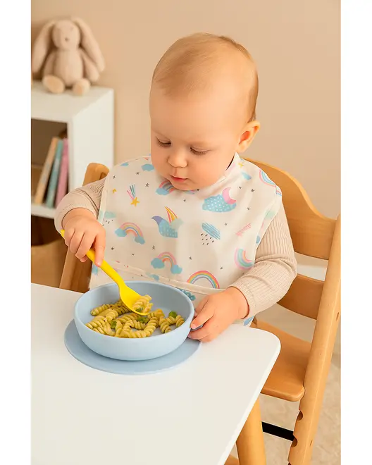 Babyhagesmæk med farverige regnbuer og skyer, vandtæt materiale, dækker bryst og skuldre, ideel til måltider. Vises på barn, der sidder ved bord og spiser pasta.